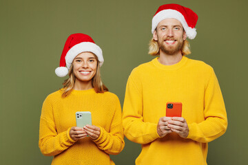 Merry smiling young couple two friends man woman wearing sweater Santa hat posing hold use mobile cell phone isolated on plain green background. Happy New Year celebration Christmas holiday concept.