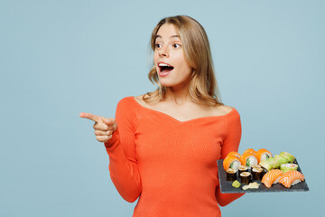 Young surprised amazed woman wear orange casual clothes point finger aside on area hold eat raw fresh sushi roll served on black plate Japanese food isolated on plain blue background studio portrait.