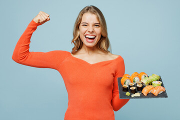 Young fun woman wear orange casual clothes show biceps muscles on hand demonstrating strength power hold eat raw fresh sushi roll served on black plate Japanese food isolated on plain blue background.