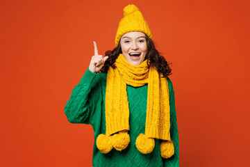 Young smiling excited happy woman she wear green knitted sweater yellow hat scarf point index finger up with great new idea isolated on plain orange red background studio portrait. Lifestyle concept.