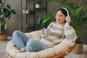 Obraz premium Side view young woman wears casual clothes listen to music in headphones drink tea sits on armchair stay at home hotel flat rest relax spend free spare time in grey living room indoor. Lounge concept.