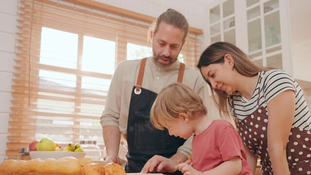 Happy family consists of father mother and little son kneading dough together in the kitchen. Caucasian parents teaching son to prepare dough for cookies and enjoying spending free time in kitchen.