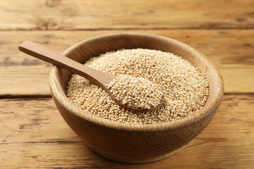 Dry quinoa seeds and spoon in bowl on wooden table, closeup