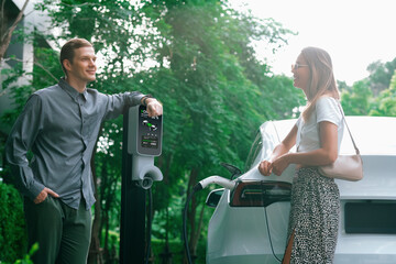 Young couple travel with EV electric car charging in green sustainable city outdoor garden in...