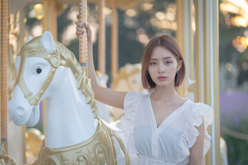 Beautiful young Asian girl in a white dress rides having fun on carnival Carousel on a horse in an Amusement Park. Portrait beauty fashion.