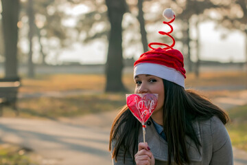 Let me love you. Beautiful young woman in grey coat and santa claus hat in park holding a lollipop in the shape of heart. Love in nature.