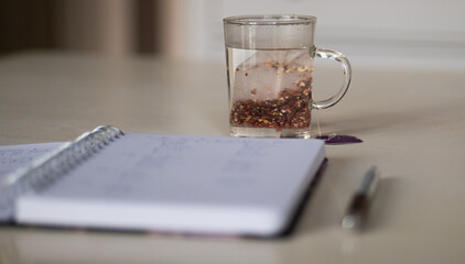 Tisane au cassis qui infuse dans une tasse transparente sur une table blanche avec un cahier et un...
