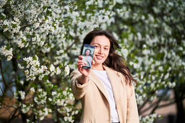 Smiling woman taking selfie with smartphone among spring blossoms with soft focus on the background.