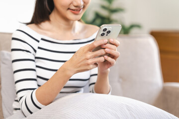 young Asian woman confidently using a smartphone, texting, chat with friends and video call sitting on a beige couch with a white cushion.