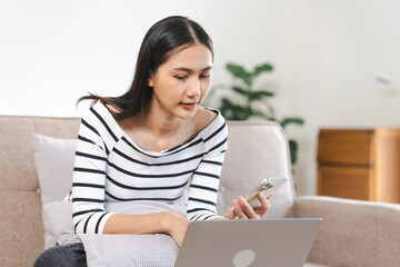 young attractive Asian woman using a laptop with a smile, sitting on a couch with a striped cushion. video call with friends or chat message