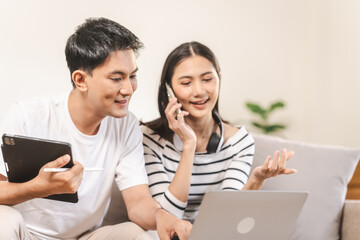 young Asian couple engaged in online shopping, wife woman holds notepad note list while husband man compares choices on smartphone. and call for price and discount, promotion.