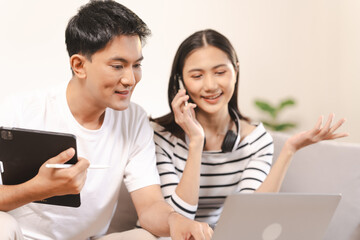 young Asian couple engaged in online shopping, wife woman holds notepad note list while husband man compares choices on smartphone. and call for price and discount, promotion.