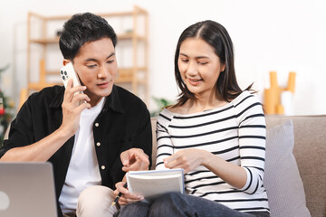 young Asian couple engaged in online shopping, wife woman holds notepad note list while husband man compares choices on smartphone. and call for price and discount, promotion.