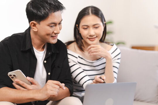 young Asian couple engaged in online shopping, wife woman holds notepad note list while husband man compares choices on smartphone. and call for price and discount, promotion.