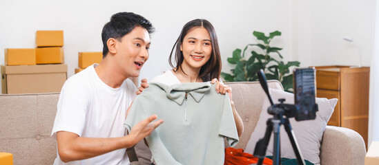 Asian people couple man and woman happily displaying products to smartphone camera for live stream in living room with boxes, home office.