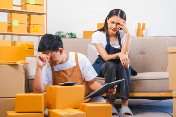 Asian man and woman look stressed, surrounded by boxes; the man holds a clipboard while the woman...