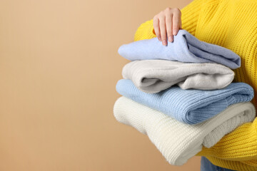 Woman with stack of casual sweaters on light brown background, closeup. Space for text - obrazy, fototapety, plakaty
