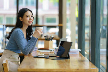 Beautiful young Asian businesswoman drinking coffee and using a laptop computer while working in an...