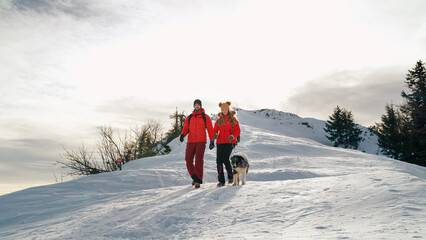 Active couple hiking on extreme terrain in the snowy mountains with their dog in winter