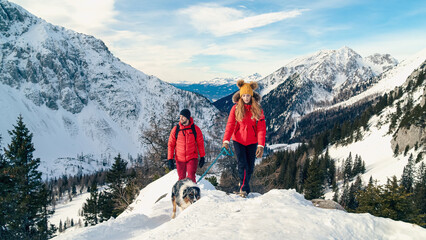 Active couple hiking on extreme terrain in the snowy mountains with their dog in winter - obrazy, fototapety, plakaty