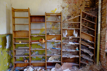 An abandoned room with a wooden bookshelf filled with old books and objects, and a crumbling brick wall. Old shelves for papers in an abandoned room. Rubbish and papers scattered around the room.