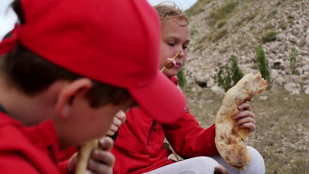 Close-up of children in red clothes on a hike eating fried sausages and fresh bread with a good voracious appetite. Teenagers on family hiking. Outdoor food cooked on fire