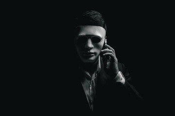 Unknown businessman wearing mask with covered face using mobile phone makes an anonymous call intimidating and threatening the interlocutor on dark background. hacker callcenter concept. - obrazy, fototapety, plakaty