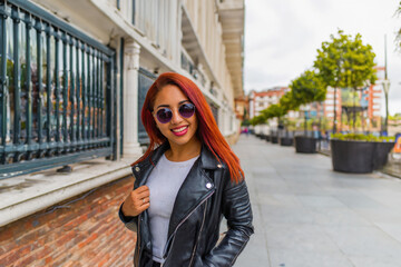  stylish young woman with red hair, wearing sunglasses and a black leather jacket, strolls on a lively urban street