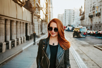 Obraz premium stylish young woman with red hair, wearing sunglasses and a black leather jacket, strolls on a lively urban street