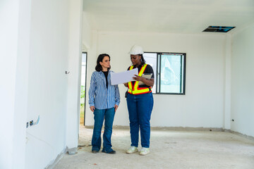 Caucasian female client inspecting house with African engineer wearing construction helmet holding blueprints for decorating, renovating a new house. Construction and Real Estate concept.