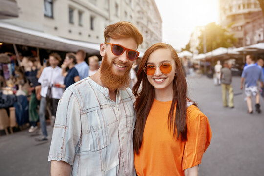 Young, bearded man and red-haired woman in orange sunglasses, smiling and posing at lively street gathering, friendly and relaxed atmosphere