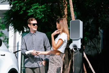 Young couple travel with EV electric car charging in green sustainable city outdoor garden in...
