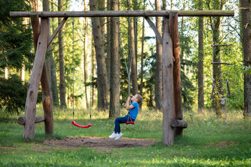 A little boy sits on a large rope swing in a sunny forest. The child plays with the ropes, twisting...