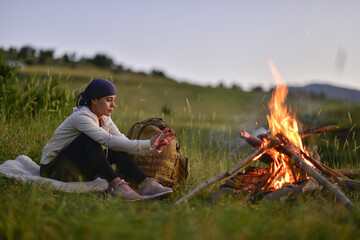 young lady camping at the foot of the mountain, watching the sunset in front of the fire - obrazy, fototapety, plakaty