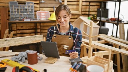 Attractive young blonde woman carpenter smiling while paying with credit card on touchpad amidst carpentry workshop - obrazy, fototapety, plakaty
