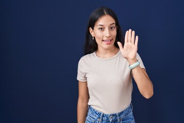 Young hispanic woman standing over blue background waiving saying hello happy and smiling, friendly welcome gesture