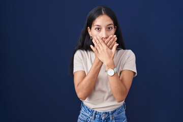 Young hispanic woman standing over blue background shocked covering mouth with hands for mistake....