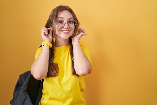 Young caucasian woman wearing student backpack over yellow background smiling pulling ears with fingers, funny gesture. audition problem