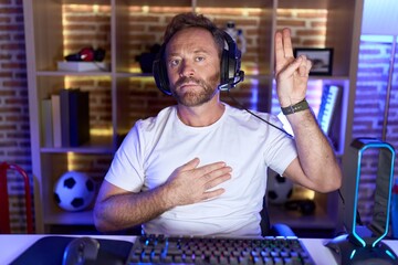 Middle age man with beard playing video games wearing headphones smiling swearing with hand on chest and fingers up, making a loyalty promise oath