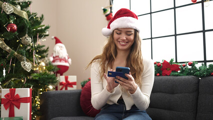 Obraz na płótnie Canvas Young beautiful hispanic woman using smartphone and credit card sitting on sofa by christmas tree at home