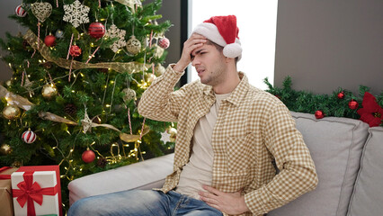 Young hispanic man wearing christmas hat having headache sitting on the sofa at home