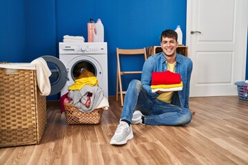 Young hispanic man holding clean laundry winking looking at the camera with sexy expression,...