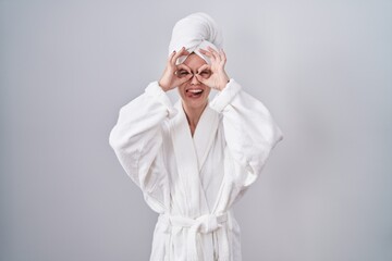 Blonde caucasian woman wearing bathrobe doing ok gesture like binoculars sticking tongue out, eyes looking through fingers. crazy expression. - obrazy, fototapety, plakaty