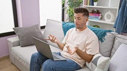 Confident young hispanic man enjoys relaxing on his comfortable sofa, using gadget at home while engaging in an exciting video call, comfortably sitting in the warm sunlight of his living room.