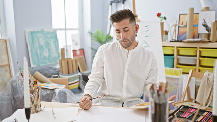 Young, handsome hispanic man ardently drawing on paper, deeply engrossed and concentrated in his...