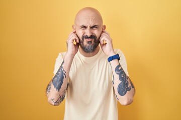 Hispanic man with tattoos standing over yellow background covering ears with fingers with annoyed expression for the noise of loud music. deaf concept.