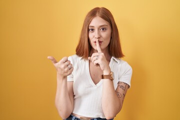 Young redhead woman standing over yellow background asking to be quiet with finger on lips pointing...