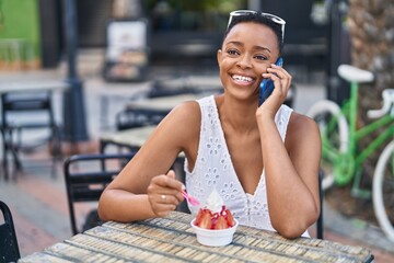 African american woman eating ice cream talking on smartphone at coffee shop terrace