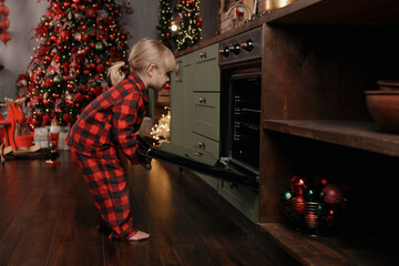 A little cute girl is standing in holiday pajamas in her home kitchen and preparing to celebrate...