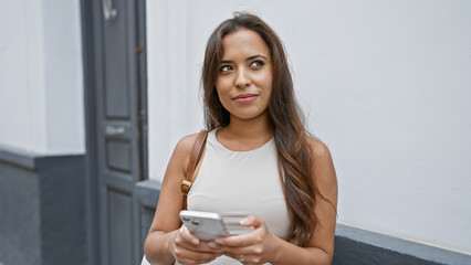 Stylish young beautiful hispanic woman standing on a sunlit city street, lost in thought, as she...